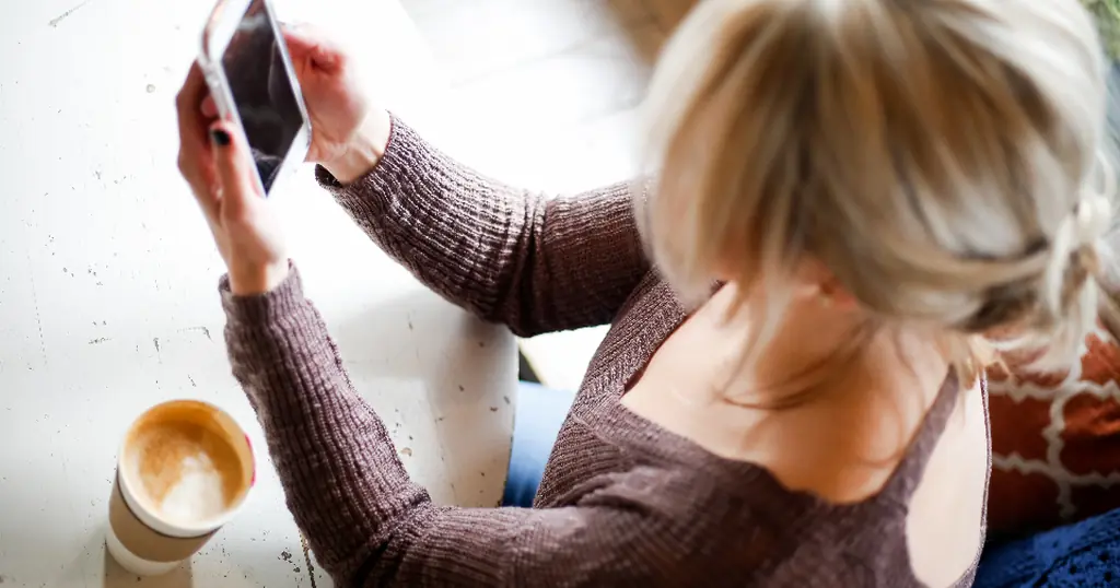 Kvinde i brun strik ved et cafébord med en kop kaffe holder mobilen op, som når man læser en første besked